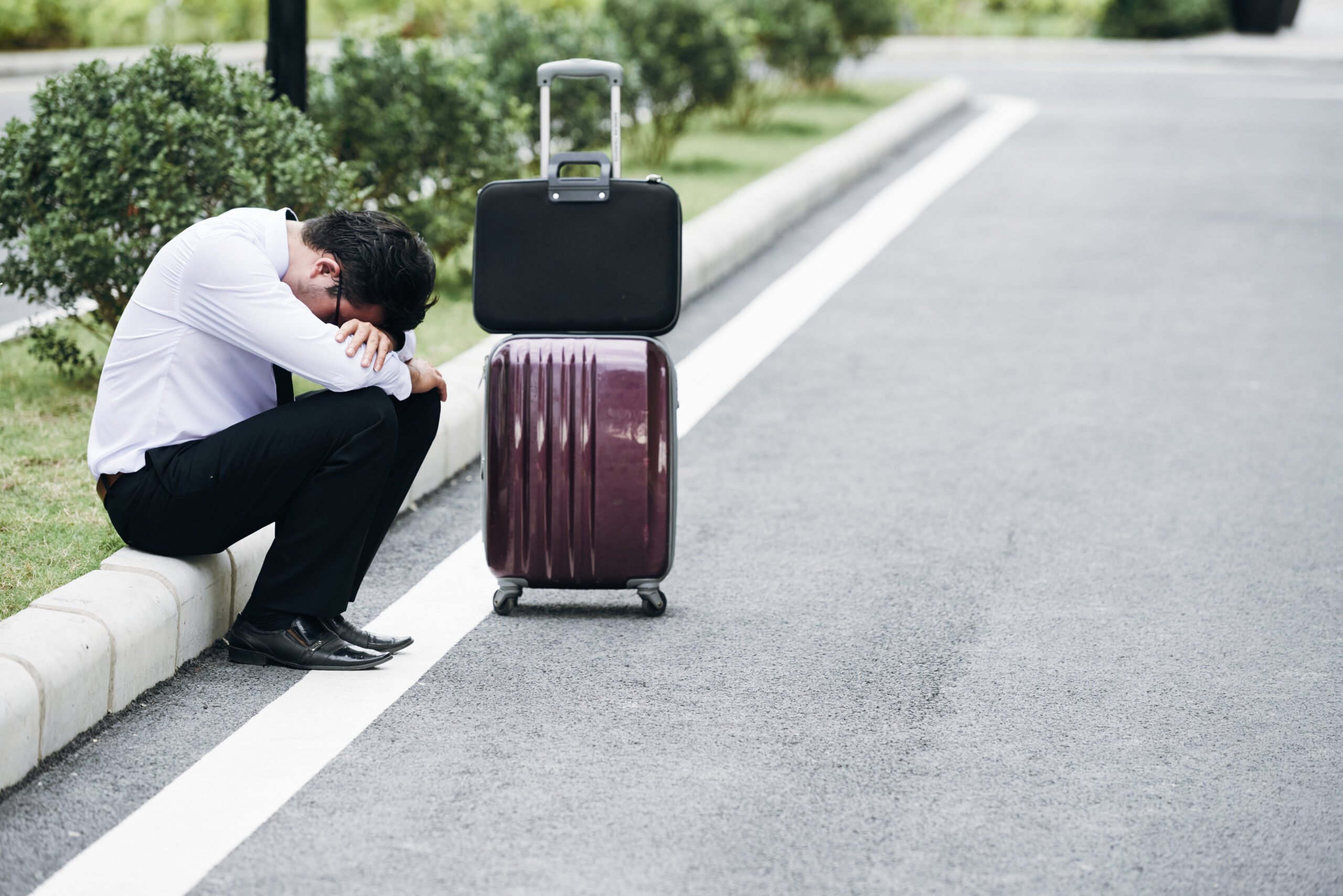 Tired or stressed businessman sitting on roadside with luggage next to him